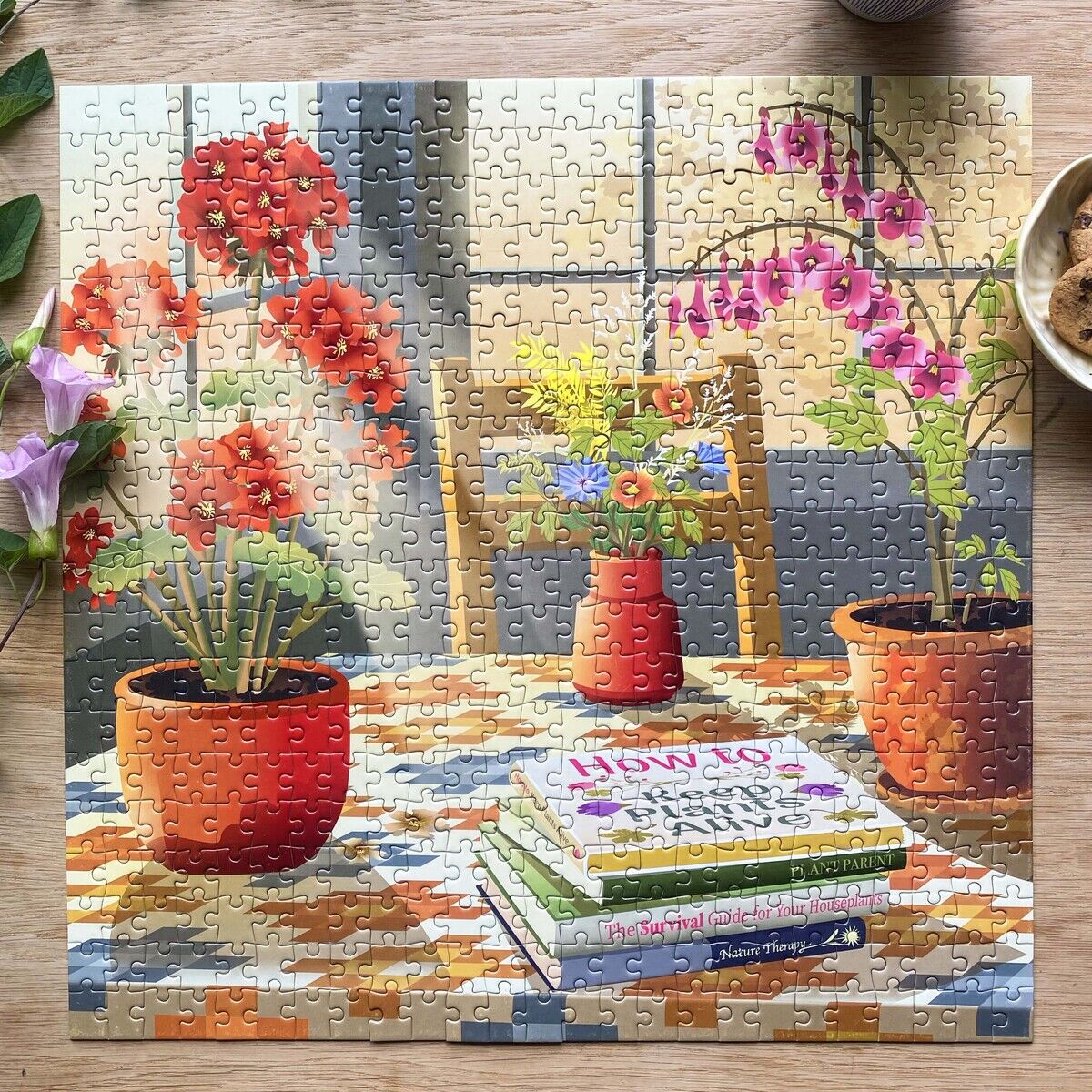 Das 500 Teile Puzzle »Blossom« von VISSEVASSE zeigt einen Holztisch mit Blumen in roten und orangefarbenen Töpfen, einem Blumenstrauß in einer Vase, einigen übereinanderliegenden Büchern und einem hellen Fenster im Hintergrund. Draußen liegen Kaffeetasse und Müslischale.