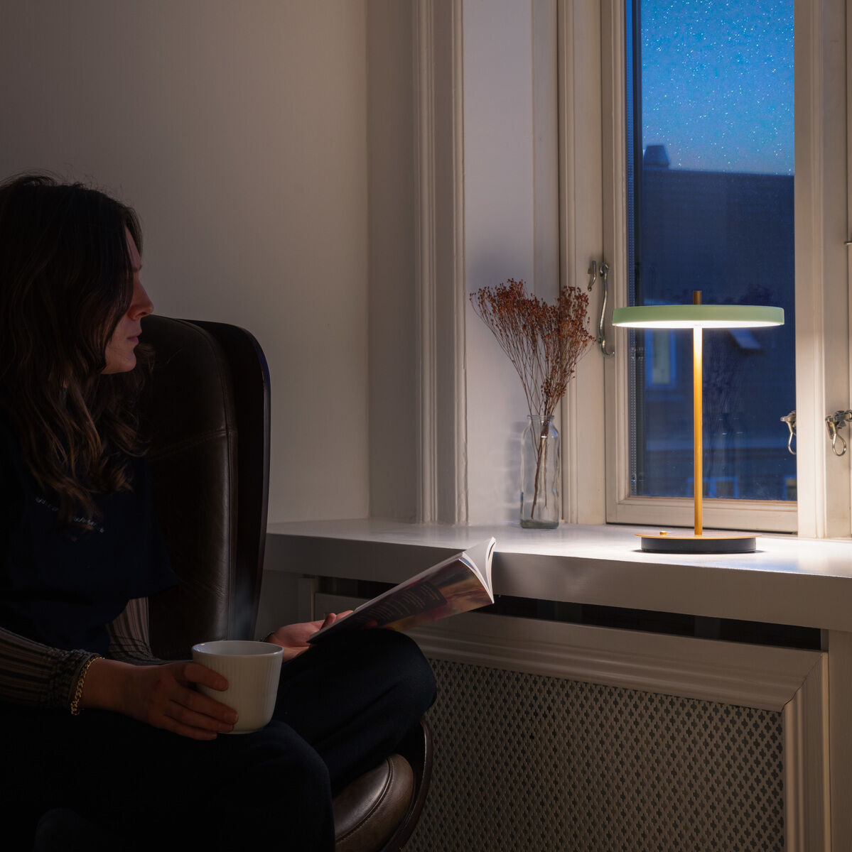 Eine moderne Tischlampe Asteria Move II steht auf einer Fensterbank und strahlt warmes Licht. Eine Person sitzt auf einem Stuhl daneben, trinkt eine Tasse und liest in einem gemütlichen Ambiente bei Nacht.