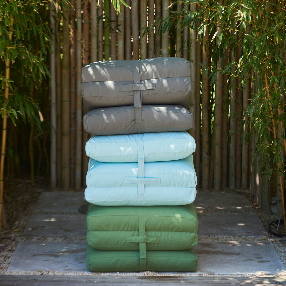 Ein Stapel von vier unterschiedlich gefärbten Gartenmatratzen »Sheffield«. Die Farben sind Grau, Hellblau und Grün. Der Stapel steht vor einem Bambuszaun und ist umgeben von grünen Pflanzen.
