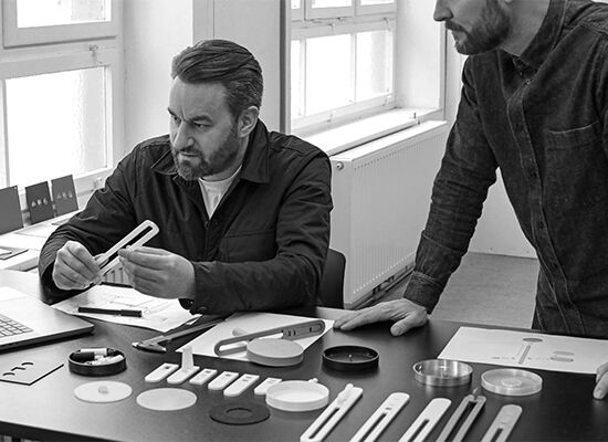 Mann mit Bart und dunkler Kleidung betrachtet ein Designobjekt am Tisch, während ein Kollege daneben steht. Auf dem Tisch liegen verschiedene Prototypen.
