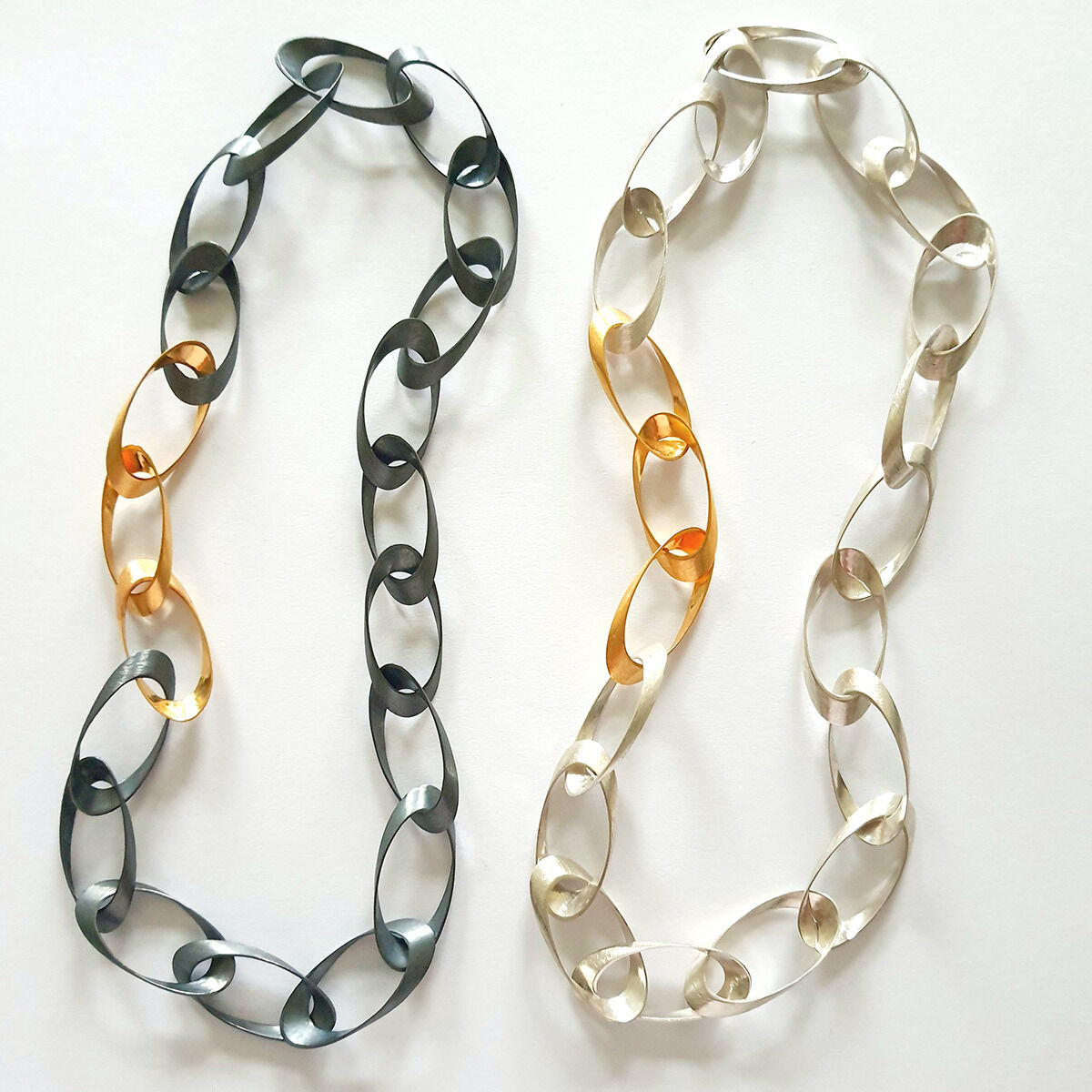 Zwei Colliers von HML Berlin, Modell „Standing Ovations“, liegen nebeneinander auf weißem Grund. Die Kette links ist teils oxidiert, teils goldplattiert, die rechte aus Silber mit goldplattierten Elementen. Die Glieder sind elliptisch, handgefertigt und glänzend.