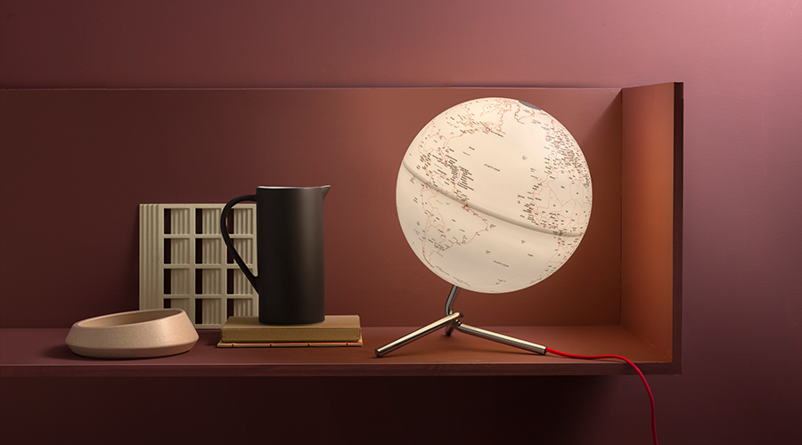Beleuchteter Globus mit weißer Oberfläche und schwarzer Schrift auf einem dreibeinigen Ständer neben schwarzem Krug, Buch und Holzschale auf braunem Regal