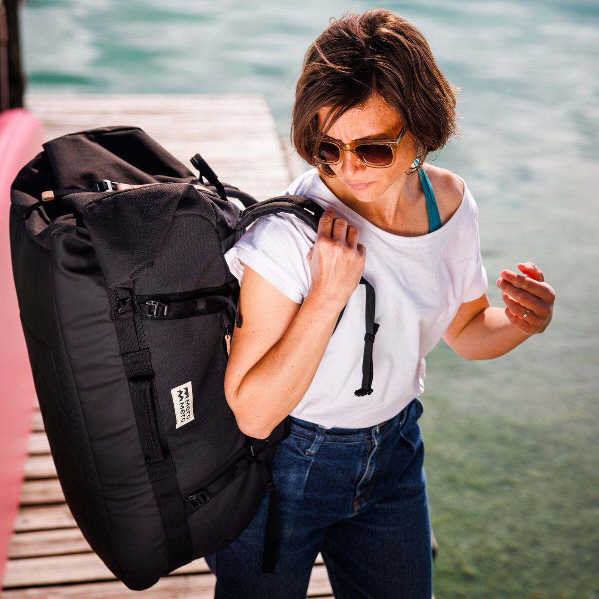 Eine Frau trägt eine schwarze Duffle-Tasche »Smögen«. Sie steht auf einem Holzsteg und trägt ein weißes T-Shirt und eine blaue Jeans. Die Umgebung deutet auf eine Outdoor- oder Freizeitaktivität hin.