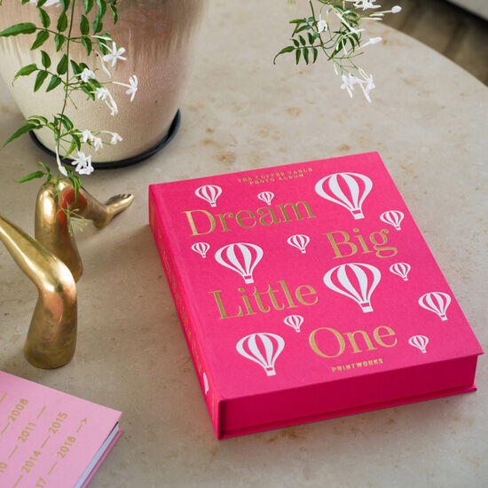 Ein rosa Babyalbum mit der Aufschrift »Dream Big Little One« liegt auf einem Tisch neben einer Vase mit Pflanzen und einer goldenen Figur.