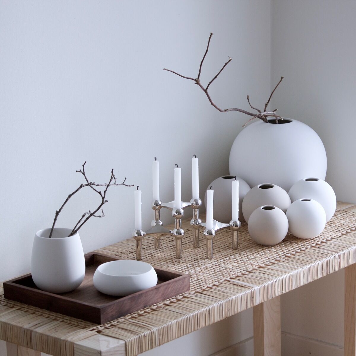 Ein moderner Kerzenständer aus Chrom steht auf einem Holztisch. Der Tisch ist mit weißen Keramikvasen und Ästen dekoriert, was ein minimalistisches und elegantes Design schafft.