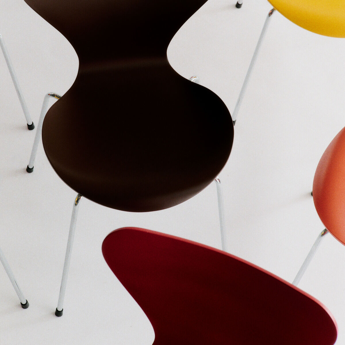 Mehrere farbenfrohe Stühle der „Serie 7“ Edition Verner Panton von Fritz Hansen stehen locker auf weißem Boden. Die Schalen sind aus lackiertem Buchenfurnier in Rot, Gelb, Orange und Dunkelbraun, die Beine aus glänzendem Stahlrohr. Modern und ikonisch.
