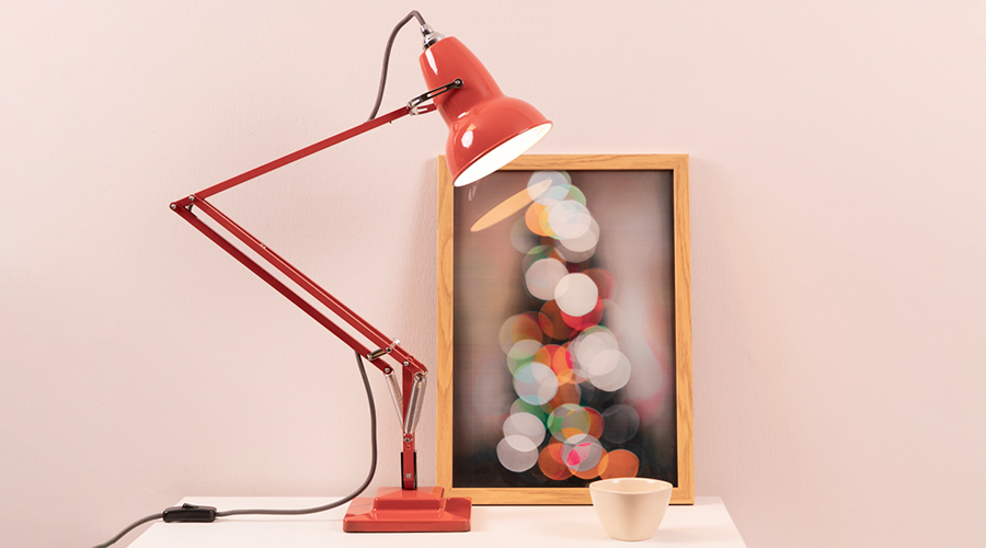 Rote Schreibtischlampe mit verstellbarem Arm neben einem Bilderrahmen mit unscharfen bunten Lichtern und einer kleinen weißen Tasse