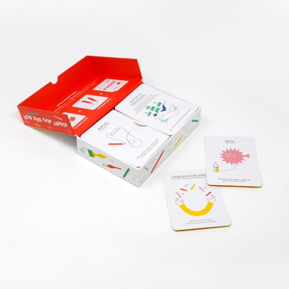 Ein Spielkartenset in einer offenen roten und weißen Schachtel. Die Karten sind illustriert und zeigen bunte Zeichnungen mit kreativen Aufgabenstellungen. Zwei Karten liegen neben der Schachtel auf weißem Hintergrund.
