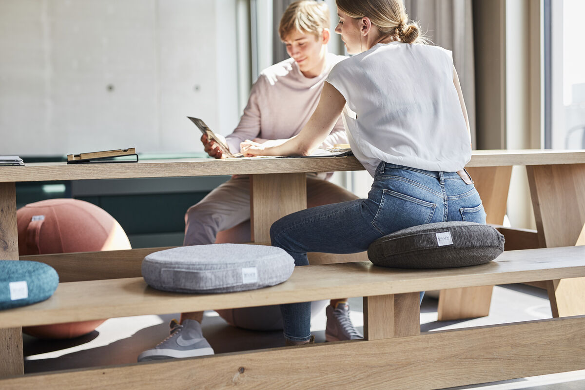 Zwei junge Menschen sitzen an einem Holztisch mit mehreren Balancekissen von VLUV. Ein Balancekissen ist grau und das andere schwarz. Im Hintergrund ist ein großes, rundes, gepolstertes Balancekissen zu sehen. Die Szene wirkt hell und modern.