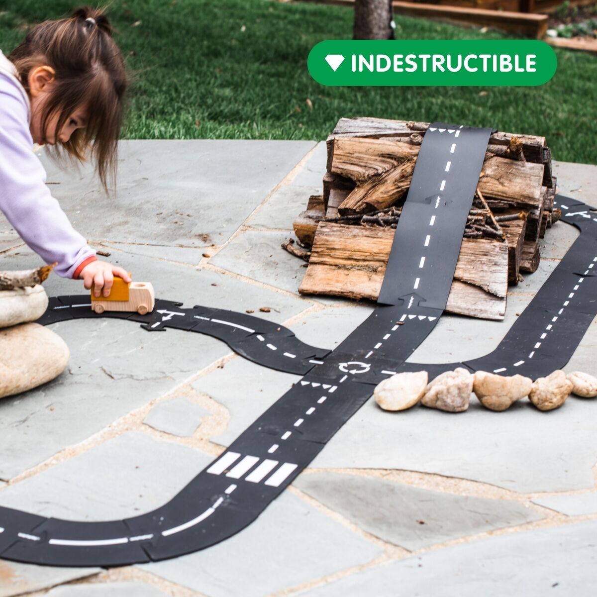 Ein Kind spielt im Freien mit einem flexiblen Straßenbau-Set auf Steinplatten. Die schwarze Spielstraße ist zu einem Straßenverlauf zusammengesetzt und wird mit Steinen und Holz ergänzt. Das Kind schiebt ein Spielzeugauto über die Piste.