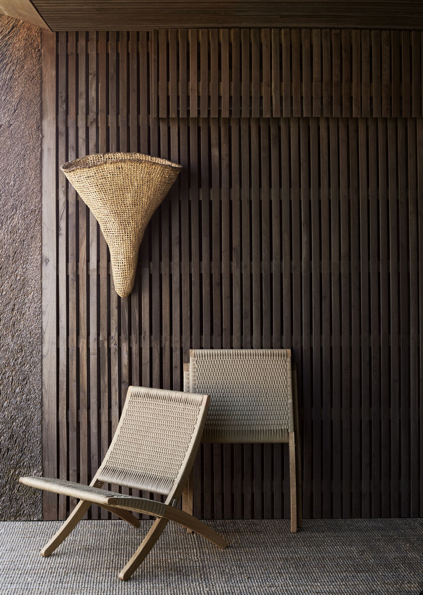 Zwei minimalistische Rattan-Stühle, die auf einem stilvollen, dunklen Holz-Hintergrund stehen. Ein gewebter Wandkorb hängt oberhalb der Stühle. Der Raum wirkt modern und naturbelassen.
