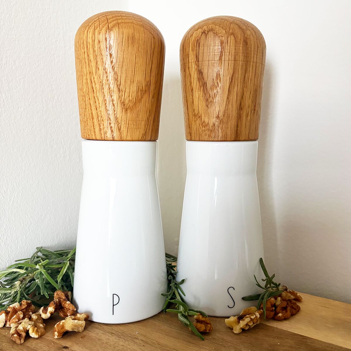 Zwei hochwertige Gewürzmühlen der ZEIT-Sonderedition »LAB« von KPM Berlin stehen nebeneinander auf einer Holzplatte. Die Mühlen bestehen aus weißem Porzellan mit gravierten Buchstaben und runden Holzaufsätzen. Zu sehen sind auch Walnüsse und frische Kräuter als Dekoration.