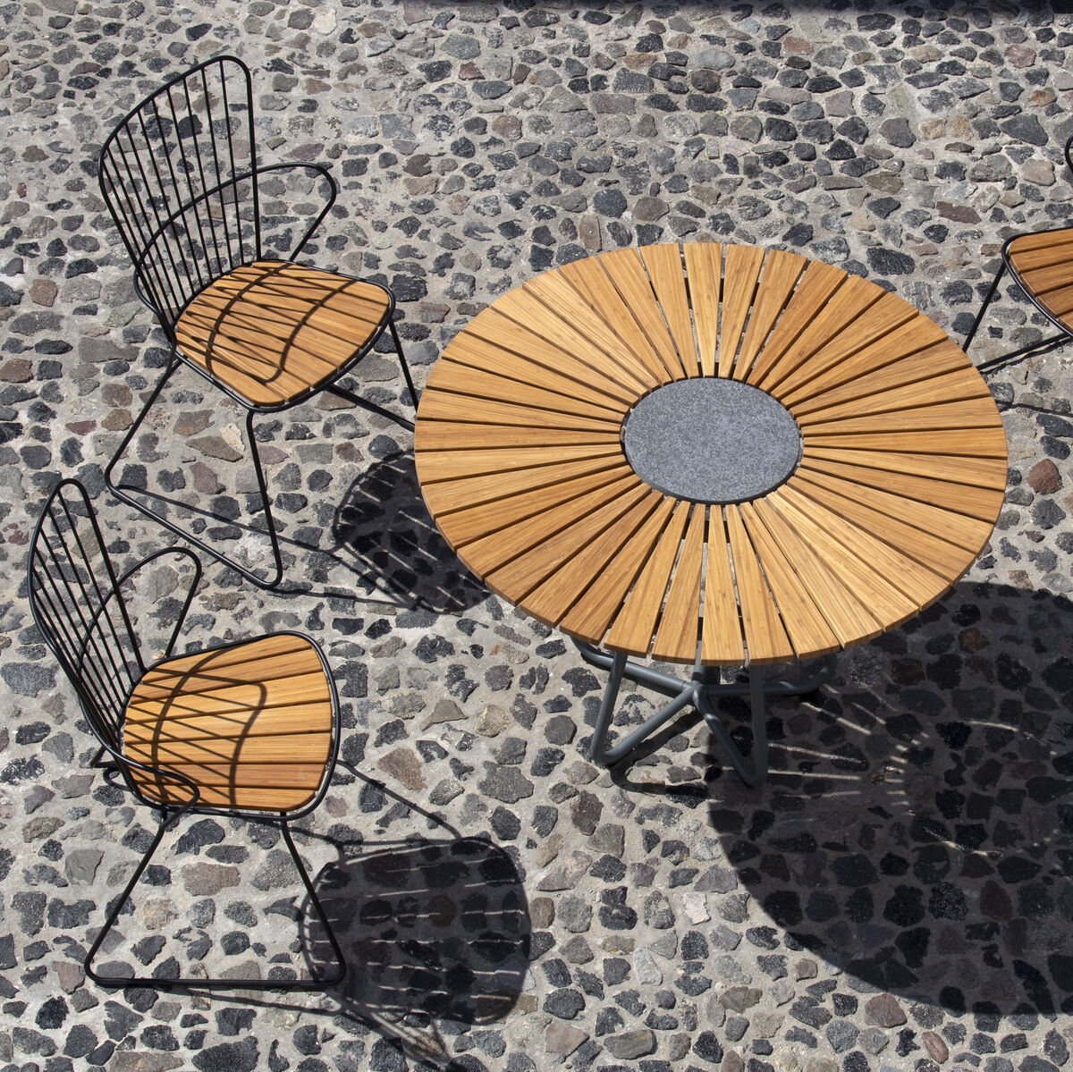 Ein runder Gartentisch mit der Bezeichnung 'Circle' aus Holz, umgeben von vier passenden Stühlen mit Metallrahmen, die auf einem gepflasterten Boden stehen.