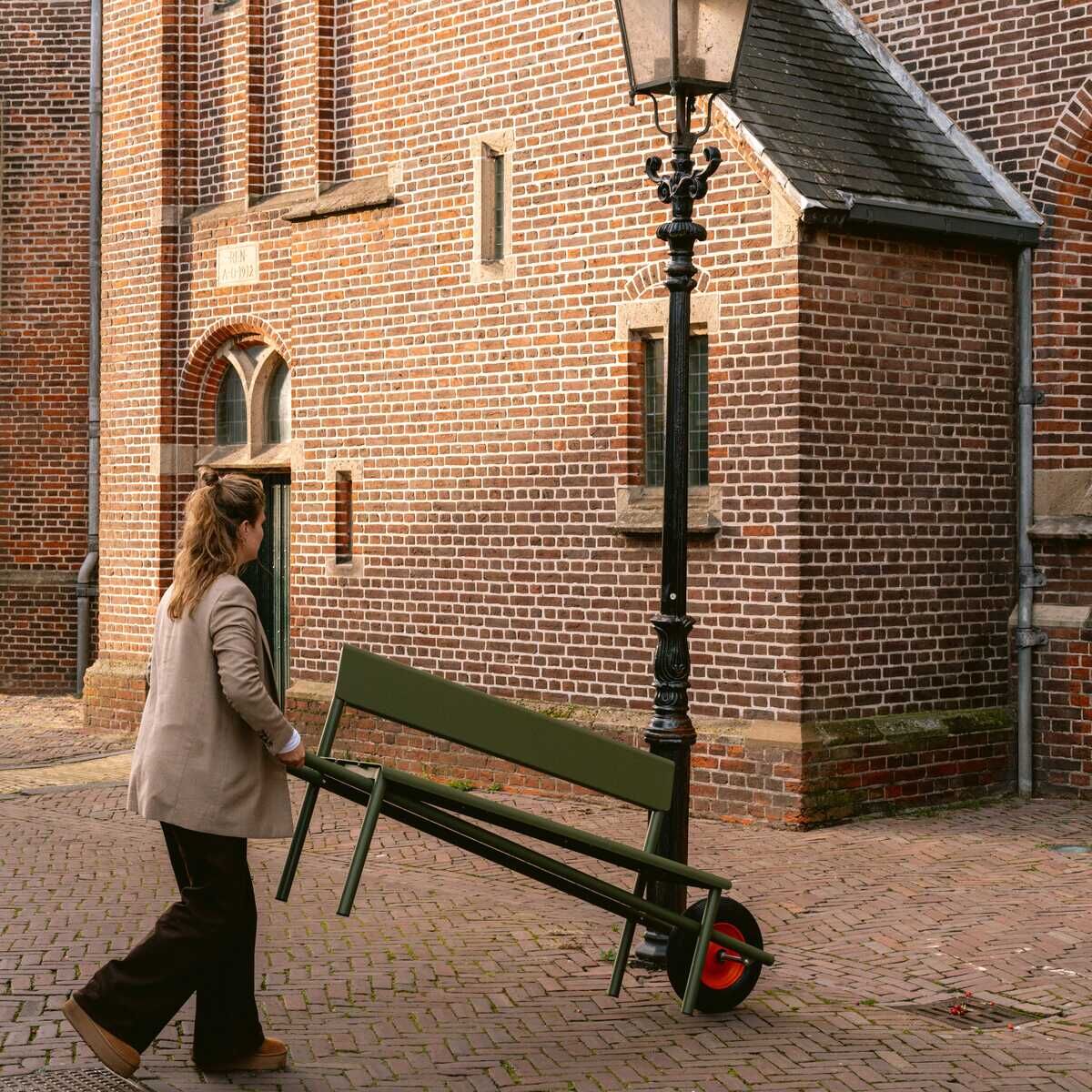 Eine Frau schiebt eine grün-dunkle Schubkarrenbank »Wheelbench« der Marke Weltevree vor einer alten Backsteinkirche entlang. Die Bank hat ein rotes Rad wie eine Schubkarre und steht auf Kopfsteinpflaster unter einer Straßenlaterne. Die Szene wirkt ruhig und urban.