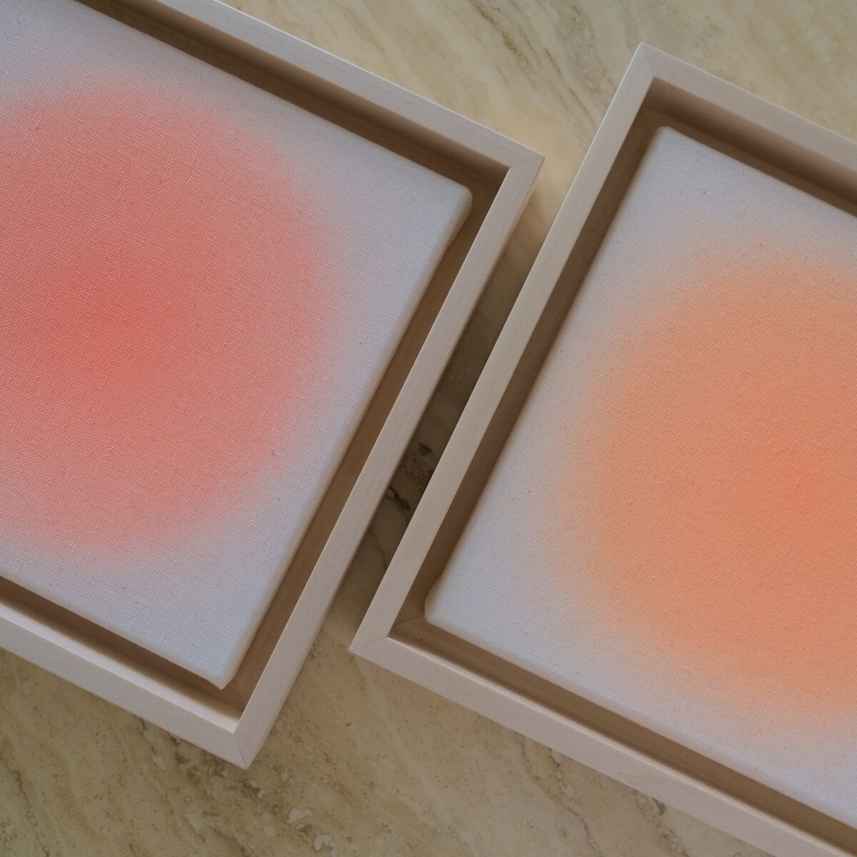 Zwei quadratische Leinwände der ZEIT-Sonderedition Sorbet »Orange« (20x20cm) auf hellem Untergrund. Beide Werke zeigen einen sanften, kreisförmigen Farbverlauf von Orange und Rosa zu Weiß, gerahmt in schlichten, hellen Holzrahmen. Die minimalistischen Bilder strahlen Wärme, Ruhe und Eleganz aus.
