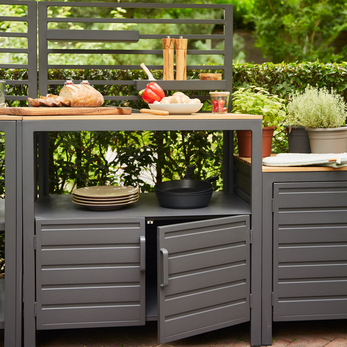 Graue modulare Outdoor-Küche auf einer Terrasse, umgeben von Pflanzen. Sie enthält Arbeitsflächen, Regale und einen Stauraum mit Türen. Auf der Arbeitsfläche befinden sich Geschirr und frisch zubereitete Lebensmittel.