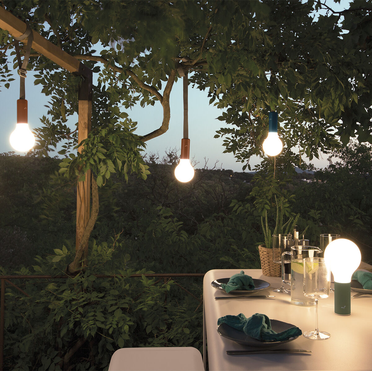 Garten mit einzeln hängenden LED-Lampen über einem Esstisch mit Gläsern und Servietten, umgeben von üppigem Grün.