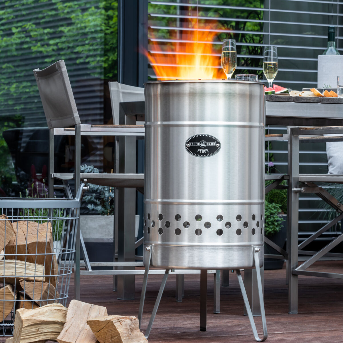 Eine moderne Edelstahl-Feuertonne Marke »Pyron« brennt auf einer Terrasse. Im Hintergrund sieht man einen gedeckten Tisch mit Speisen und Getränken sowie einige Gartenstühle. Holzscheite liegen in einem Metallkorb daneben.