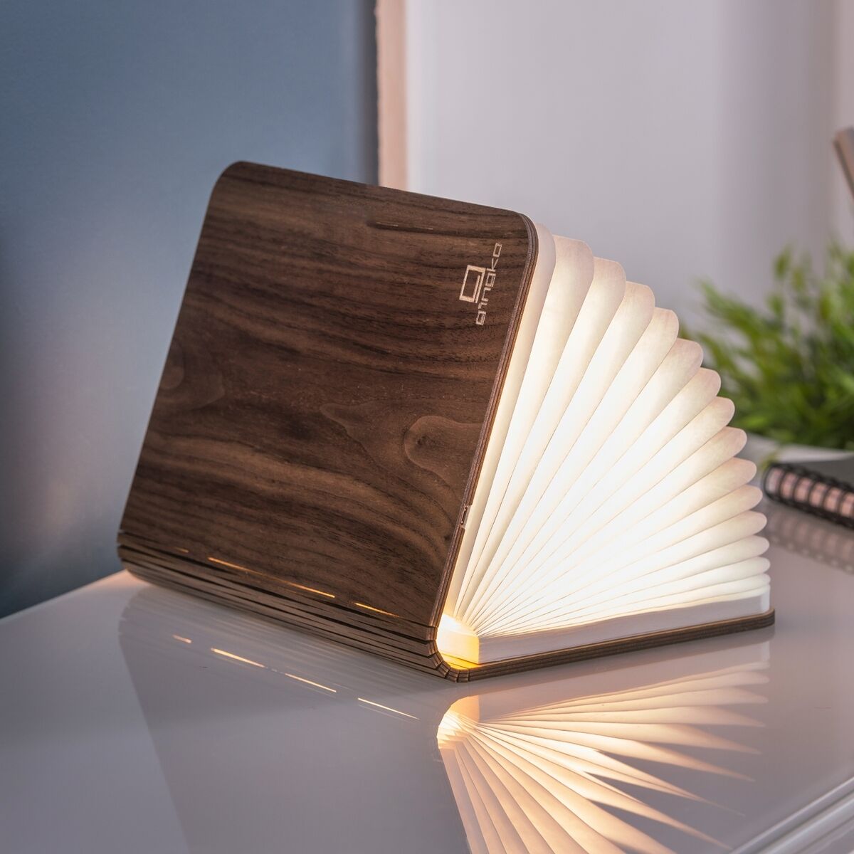 Ein Smart Booklight in Walnussholz-Optik, aufgeklappt und leuchtend, steht auf einem weißen Tisch. Im Hintergrund sind ein Stiftehalter mit Schreibutensilien, ein Notizbuch und eine Pflanze zu sehen.
