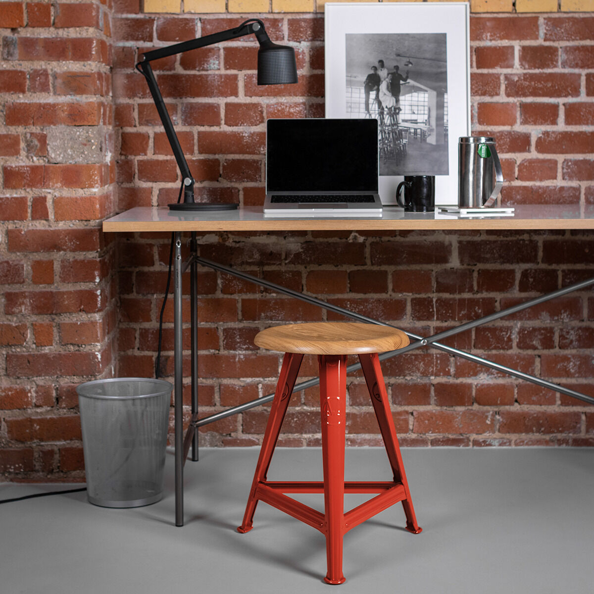 Ein industriell gestalteter Raum mit einer Ziegelsteinwand, einem Schreibtisch und einem roten Schemel. Auf dem Schreibtisch stehen eine Lampe, ein Laptop, und Dekorationselemente.