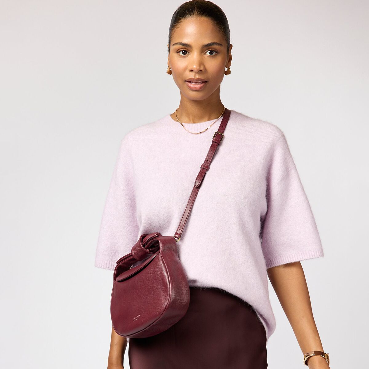 Frau mit blassrosafarbenem Strickpullover trägt eine halbmondförmige, bordeaux-dunkelrote Umhängetasche „Coco Croissant“ aus softem Narbenleder mit edlen Beschlägen aus antikem Messing. Die Tasche wirkt luxuriös und elegant, abgestimmt auf den monochromen Look.