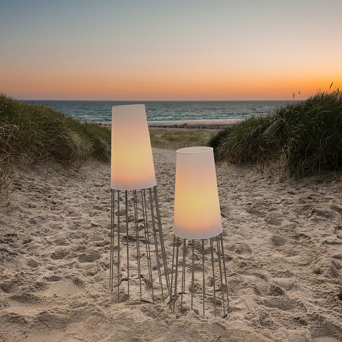 Zwei Gartenlichter »Lighthouse« stehen im Sand eines Strandwegs, der zum Meer führt. Die Lampen sind auf hohen Metallbeinen und leuchten sanft in der Abenddämmerung mit einem Sonnenuntergang im Hintergrund.