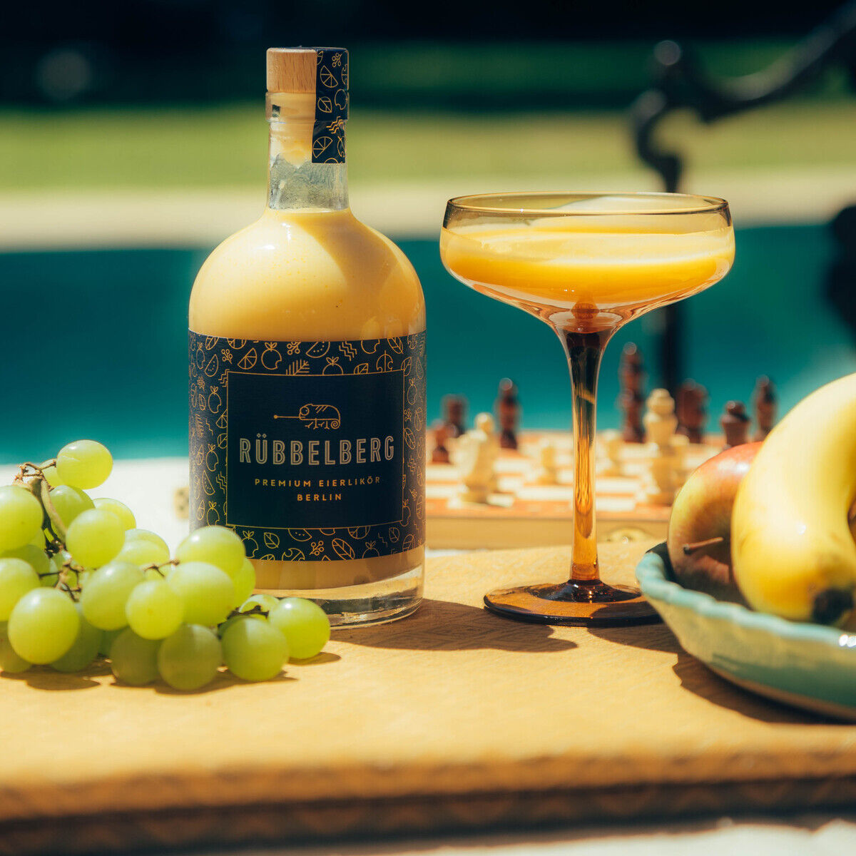 Eine 500ml Flasche Premium Eierlikör der Marke Rübbelsberg steht auf einem Holztisch neben einem gefüllten Likörglas, frischen Weintrauben und Obst im Sonnenschein. Im Hintergrund ist ein Swimmingpool und eine Schachfigur zu sehen. Das Bild vermittelt sommerliche und genussvolle Stimmung.