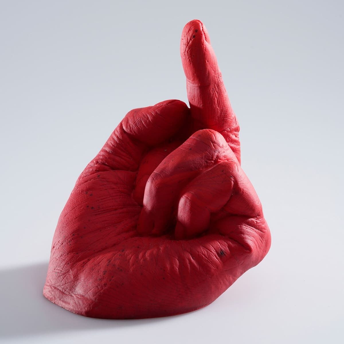 Eine rote, glasartige Plastikskulptur in Form einer Hand mit ausgestrecktem Mittelfinger auf einem weißen Hintergrund. Das Kunstwerk trägt den Titel 'Study of Perspective in Glass' von Ai Weiwei.