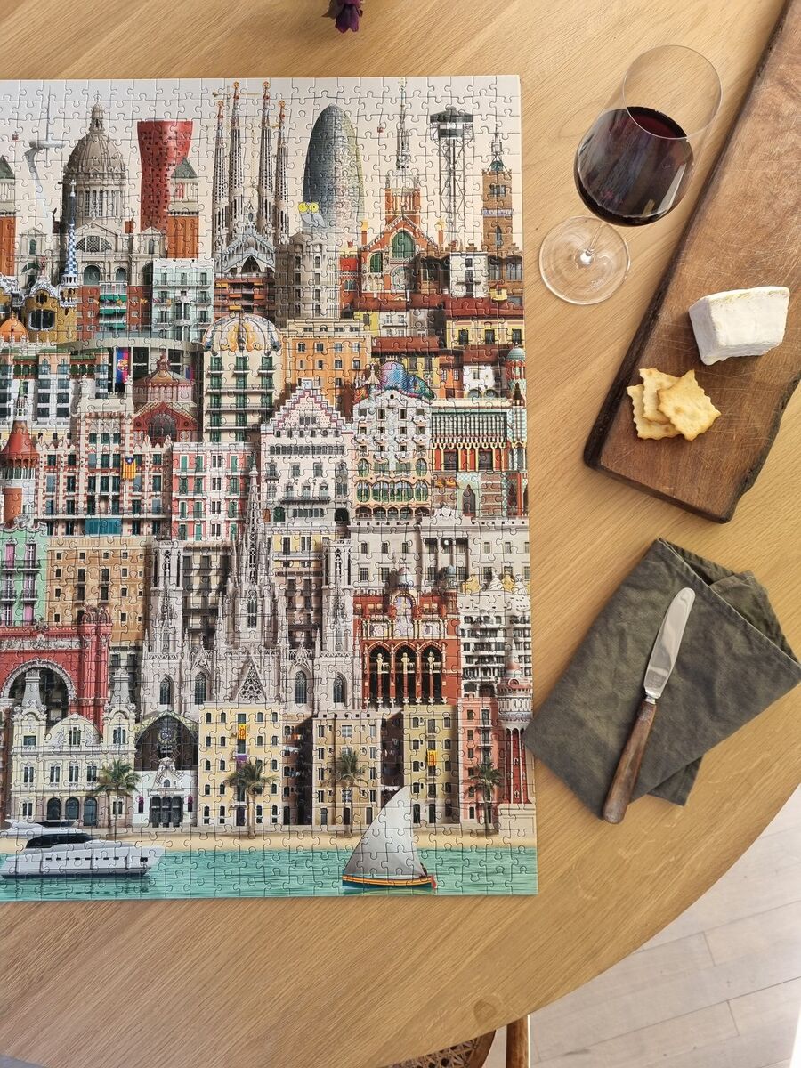 Ein 1000-Teile-Puzzle mit einer detaillierten Darstellung von Barcelonas Architektur, inklusive berühmter Gebäude wie der Sagrada Familia. Im Vordergrund sind ein Glas Rotwein, Käse und Cracker auf einem Holzbrett sowie ein Messer und eine grüne Serviette zu sehen, die auf einem Holztisch liegen.