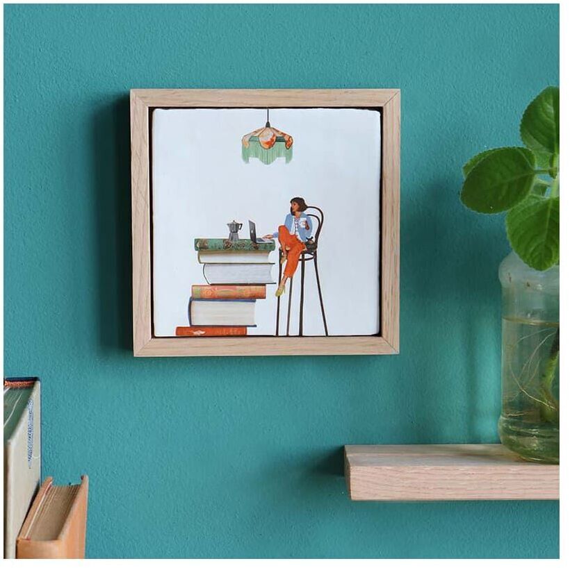 Eine Keramikfliese mit Holzrahmen zeigt eine Illustration eines Menschen, der auf einem Stapel Bücher sitzt und liest. Die Fliese hängt an einer grünen Wand und ist neben einem Bücherregal und einer Pflanze platziert.