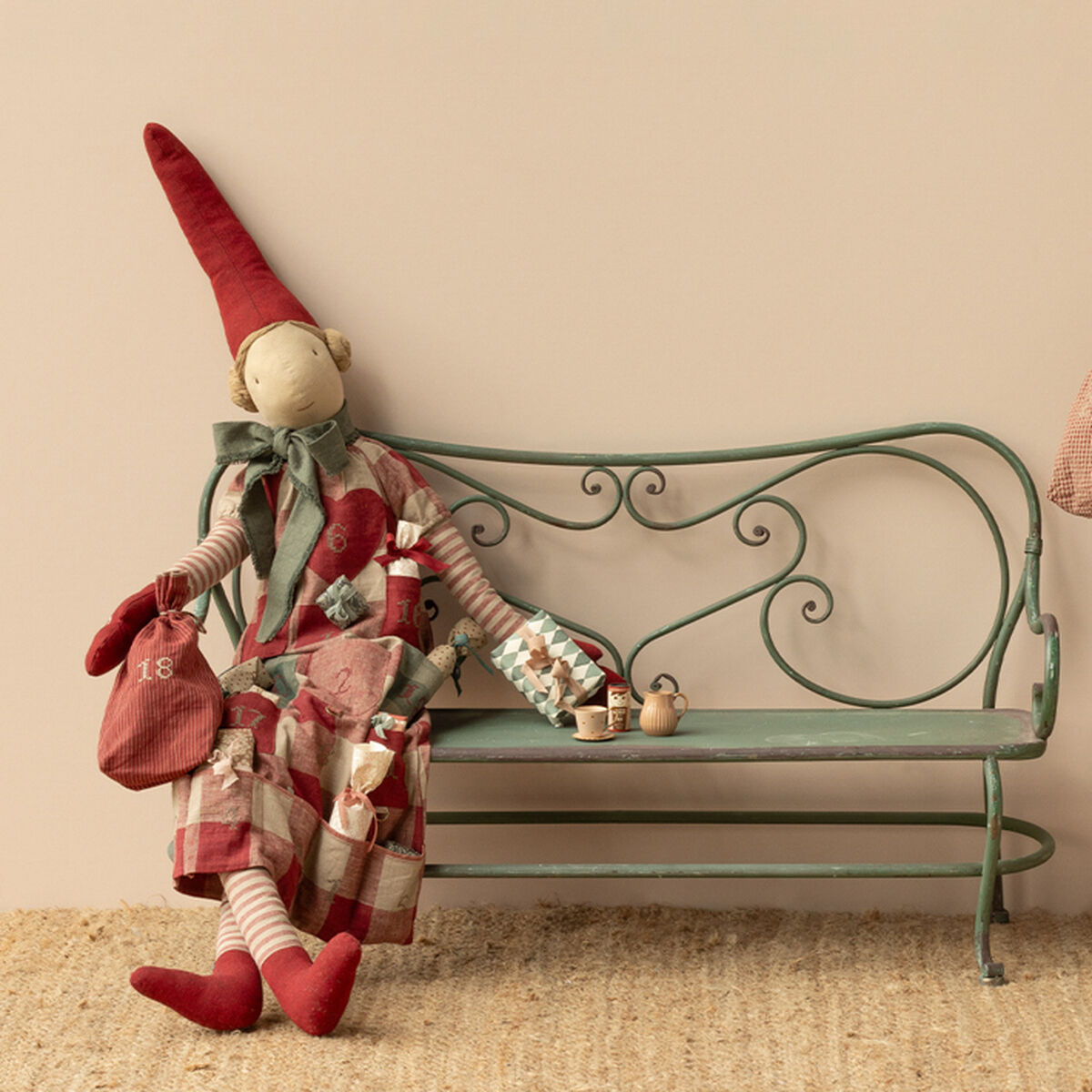 Zwei Stoffpuppen in Form von Pixy Mädchen mit roten Zipfelmützen und gestreiften Kleidungsstücken stehen neben einer dekorativen Metallbank und einem kleinen Tannenbaum. Die Szene ist weihnachtlich dekoriert und die Figuren tragen kleine Geschenke sowie Adventssäckchen. Der Hintergrund ist neutral gestaltet.