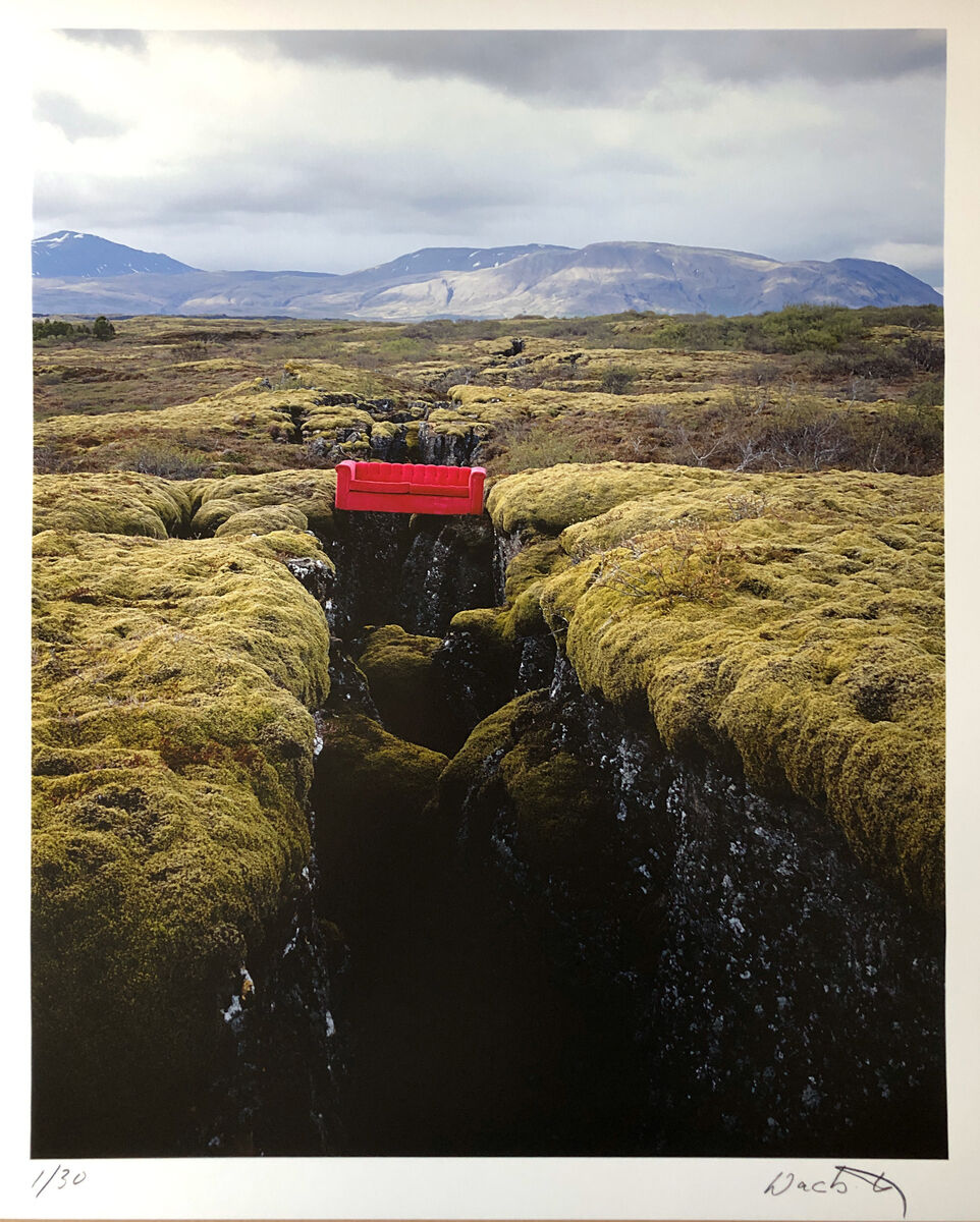 Fotografie einer moosbedeckten Landschaft in Island. In der Mitte verläuft ein Spalt in den Felsen, über dem eine rote Bank steht. Der Himmel ist bewölkt und im Hintergrund sind Berge zu sehen.
