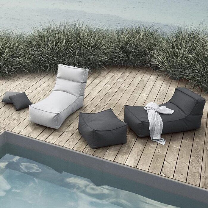 Outdoor-Lounge-Bereich mit modernen grauen und weißen Loungemöbeln auf einer Holzterrasse neben einem Pool. Gräser säumen den Hintergrund am Wasser.