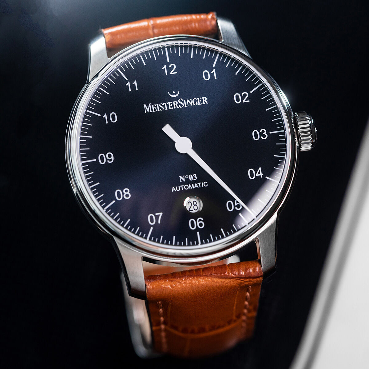 Die ZEIT-Sonderedition »N°03« von MeisterSinger ist eine elegante Armbanduhr mit einem schwarzen Zifferblatt und einem einzigen Stundenzeiger. Sie hat ein Edelstahlgehäuse und ein braunes Lederarmband, sowie eine Datumsanzeige bei sechs Uhr.