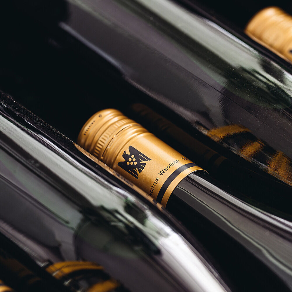 Nahaufnahme mehrerer liegender Weinflaschen der Weinedition »Beethoven« von Weingüter Wegeler mit goldfarbenen Schraubverschlüssen, auf denen das Logo und Markenname sichtbar sind. Die Flaschen spiegeln ein elegantes, hochwertiges und exklusives Ambiente wider.
