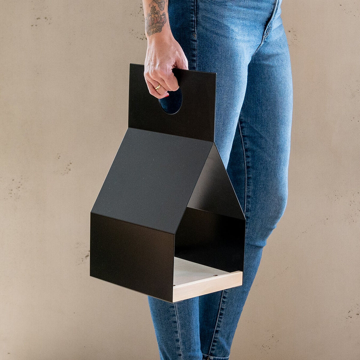 Eine Person in blauer Jeans und weißen Sneakern hält das moderne, schwarze Vogelhaus »FLY« der Marke finbeaux von Christoph Schoen in der Hand. Das Vogelhaus hat eine geometrische Form, einen Tragegriff und steht auf hellem Holzboden vor neutraler Wand.
