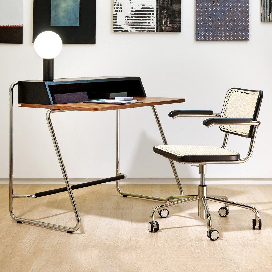 Schreibtischstuhl »S 64 Atelier« von Thonet: Ein stilvoller Bürostuhl mit schwarzem Sitzkissen und Lehne, kombiniert mit einem klassischen Design aus Stahlrohrrahmen. Der Stuhl hat Rollen und ist höhenverstellbar. Im Hintergrund ist ein moderner Schreibtisch mit verschiedenen Büchern und Dekorationsgegenständen zusehen, eingerahmt von Kunstwerken an der Wand.
