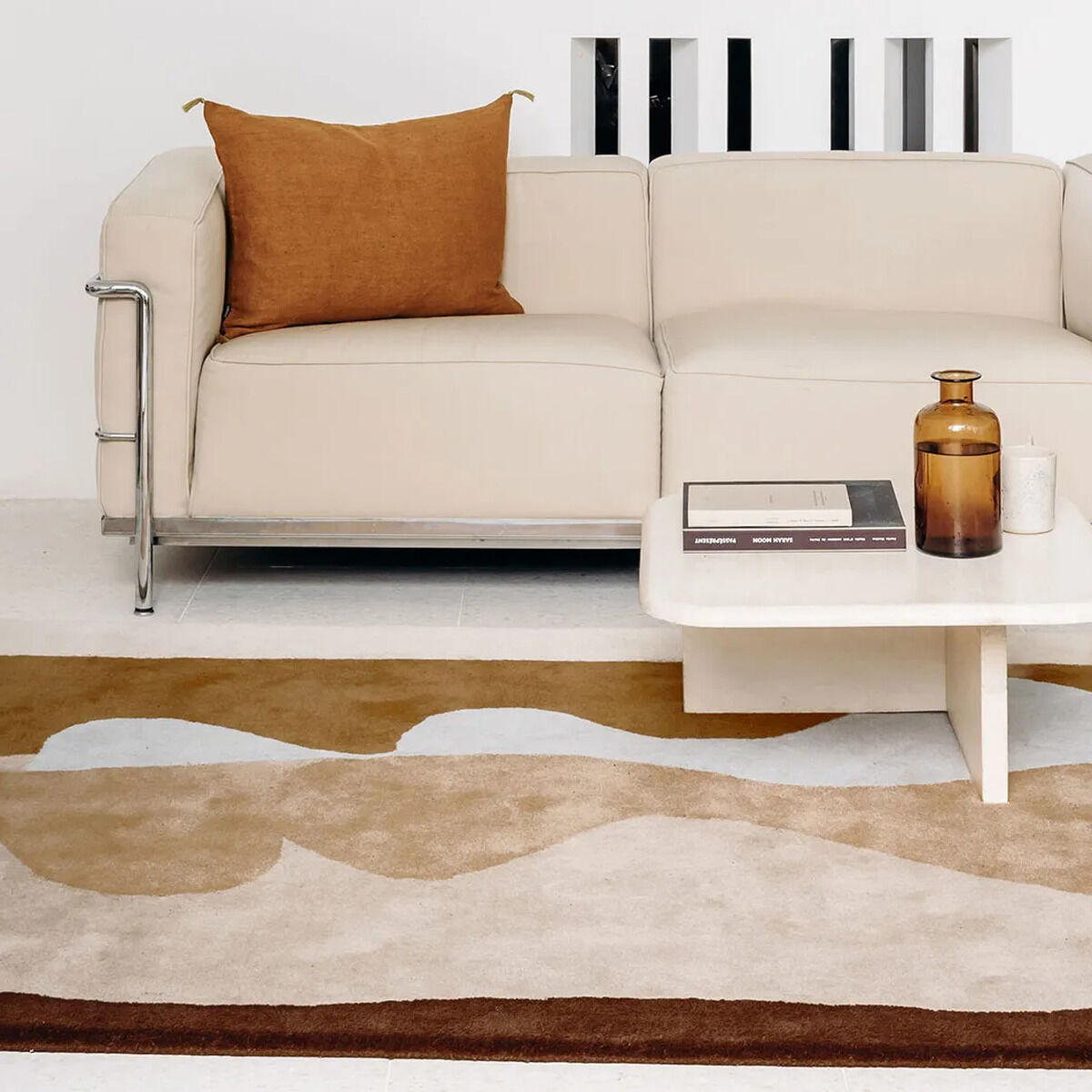 Moderner Teppich mit abstraktem Muster in Ocker-, Beige- und Brauntönen, ausgelegt vor einem hellen Sofa in einem minimalistisch eingerichteten Wohnzimmer mit kleinem weißen Couchtisch und dekorativen Elementen.