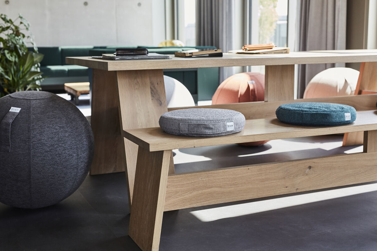 Ein modernes Wohnzimmer mit einem großen Holztisch und mehreren Balancekissen der Marke VLUV in verschiedenen Farben, darunter Grau und Grün. Die Kissen sind auf einer Holzbank platziert und im Hintergrund sind weitere ergonomische Sitzmöbel zu sehen.