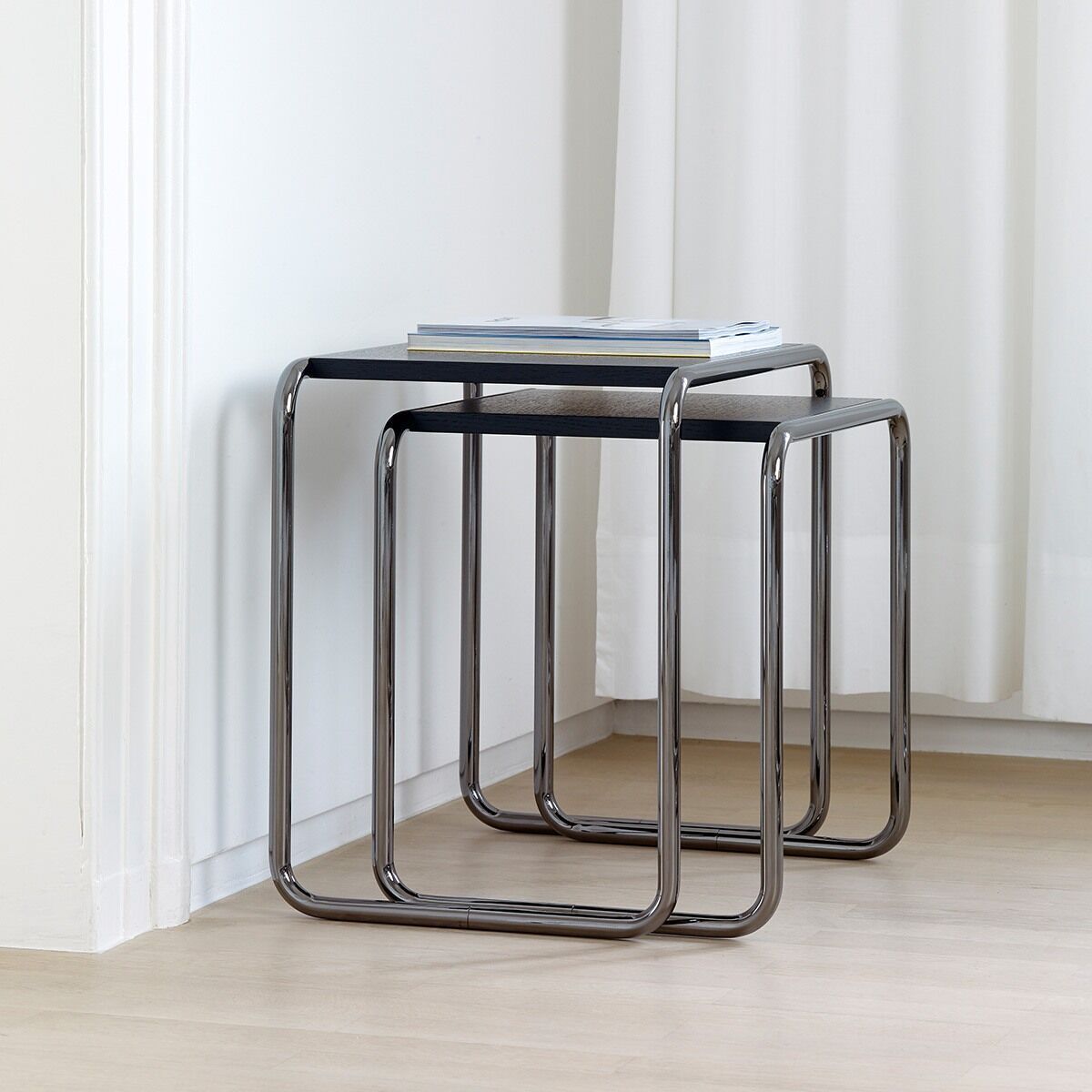Ein Beistelltischsatz der ZEIT-Sonderedition B9a/b von Thonet, bestehend aus zwei ineinander verschachtelten Tischen. Die Tische haben eine schwarze Oberfläche und ein Gestell aus poliertem Metall. Hintergrund ist ein heller Raum mit Holzboden und einem weißen Vorhang.