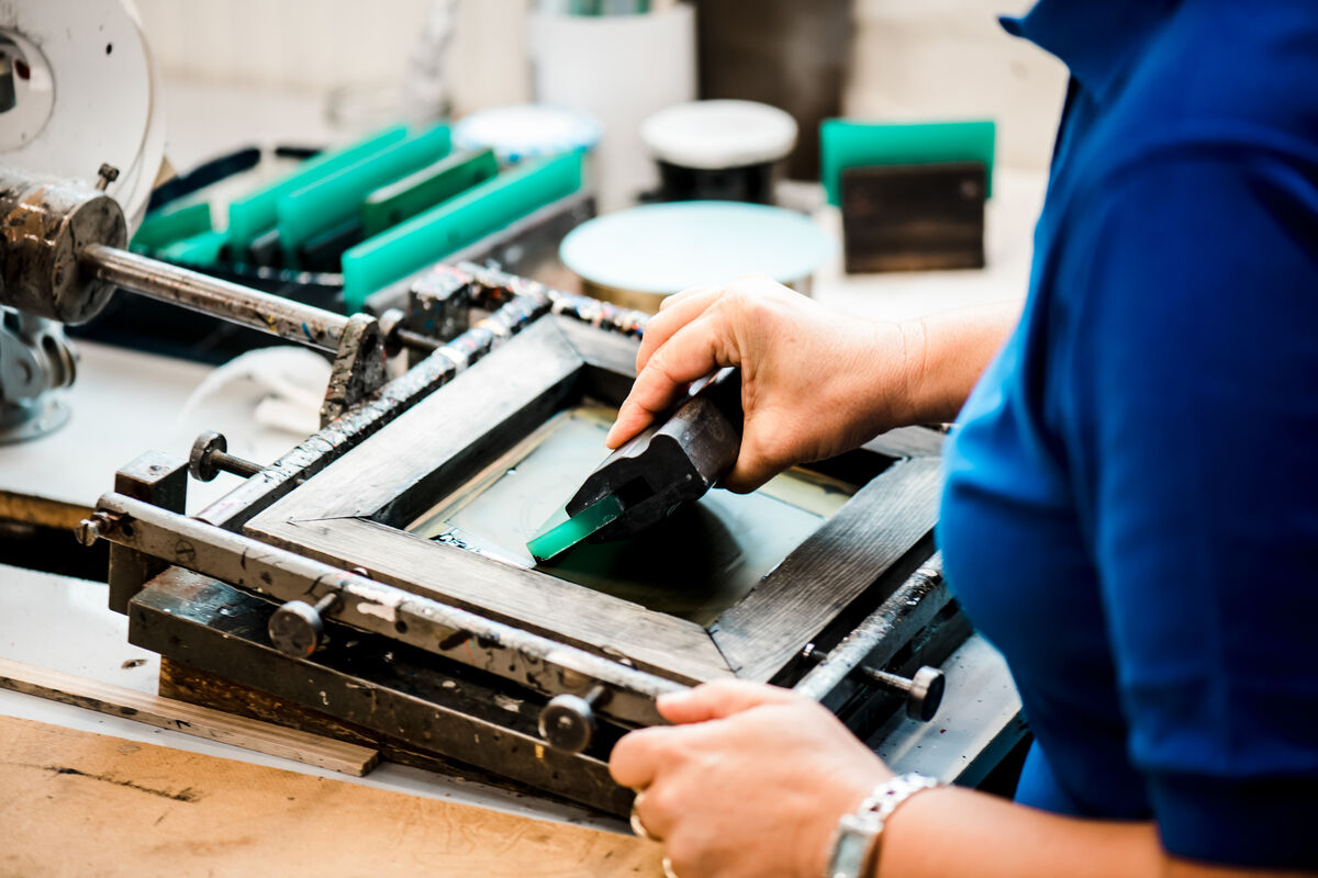 Eine Person führt präzise Handwerksarbeit an einer Maschine aus. Es sind diverse Werkzeuge und grüne Kunststoffteile sichtbar. Die Person trägt ein blaues Shirt.
