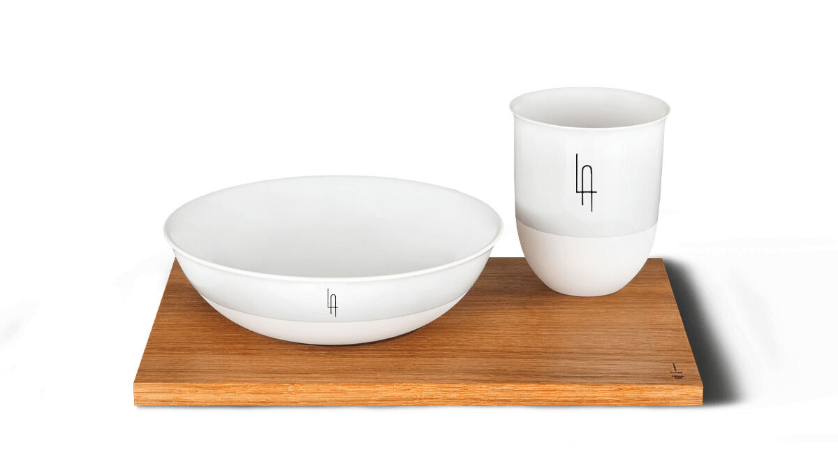 Ein Müsli-Set bestehend aus einer weißen Porzellanschale und einem weißen Porzellanbecher mit Monogramm auf einem hellbraunen Holztablett.