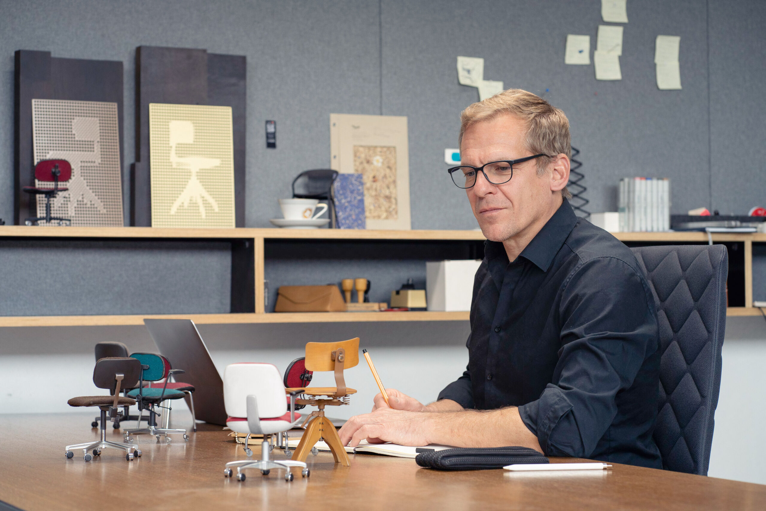 Ein Designer sitzt an einem Holztisch mit Miniaturstühlen, einem Laptop und Skizzen, umgeben von kreativen Materialien und einem organisierten Arbeitsbereich.