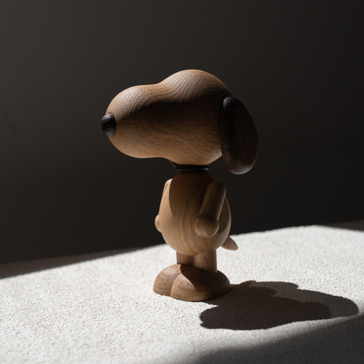 Holzfigur der Comicfigur Snoopy, in natürlicher Holzfarbe, steht in einem minimalistischen Design auf einer weißen Oberfläche. Das Licht und der Schatten betonen die Details der Figur.