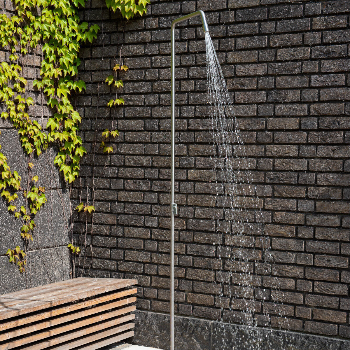 Eine Gartendusche »Serpentine« mit Edelstahldesign steht neben einer Holzbänke vor einer Backsteinmauer, die mit grünen Pflanzen bewachsen ist. Wasserstrahlen fließen aus der Duschnopf.