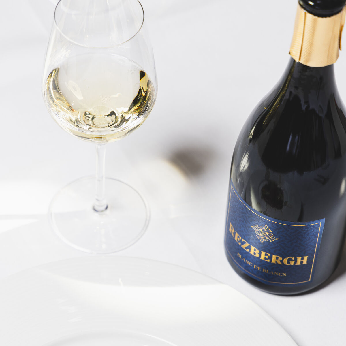 Auf einem weiß gedeckten Tisch stehen eine dunkelblaue Flasche Crémant de Luxembourg und ein gefülltes Sektglas. Daneben liegt ein edel angerichtetes Carpaccio mit Käse, grünen Kräutern und kleinen Brotwürfeln auf einem weißen Teller.