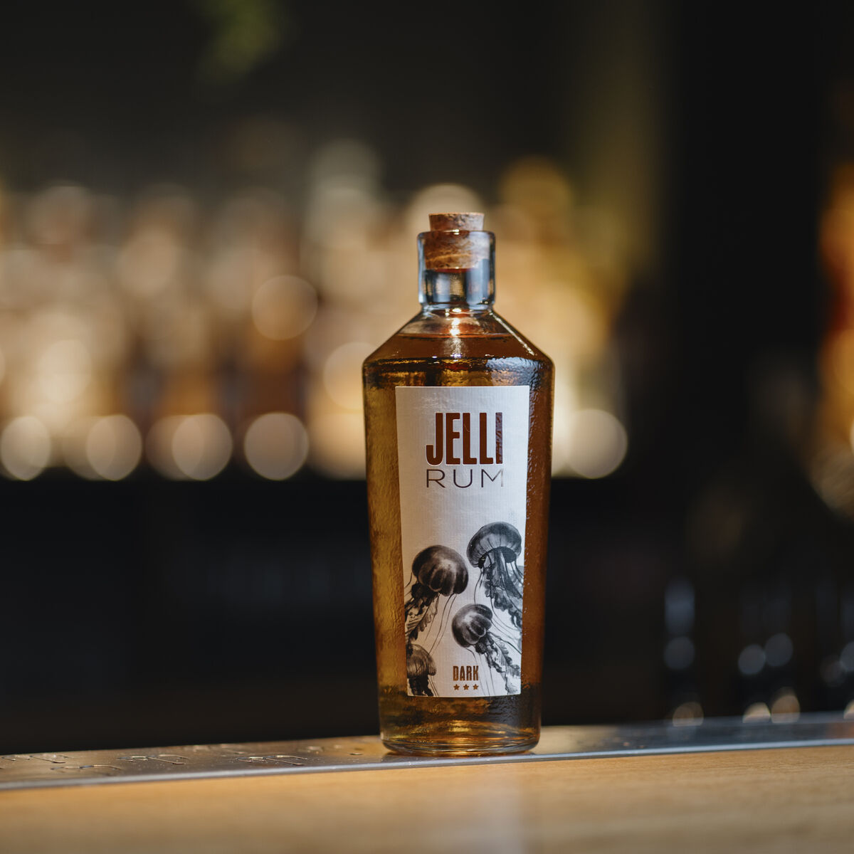 Die Abbildung zeigt eine Flasche JELLI Rum Dark auf einer Holzoberfläche vor unscharfem Barhintergrund. Das Etikett ist dekorativ gestaltet, die Flasche ist eckig mit Schraubverschluss und enthält eine dunkelbraune Flüssigkeit. Im Hintergrund sind warme Lichtquellen sichtbar.