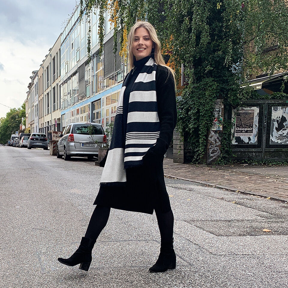 Eine Frau steht auf einer städtischen Straße und trägt einen schwarz-weiß gestreiften Seidenschal. Im Hintergrund sind geparkte Autos und Gebäude zu sehen.