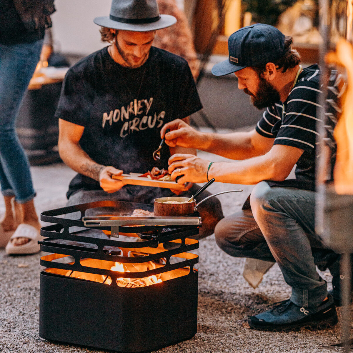 Zwei Personen sitzen um eine moderne schwarze Feuerstelle im Freien. Einer von ihnen grillt Essen auf der Plancha. Im Hintergrund ist ein gemütliches Ambiente mit Pflanzen und subtiler Beleuchtung zu sehen. Das Szenario unterstreicht eine gesellige und entspannte Atmosphäre.