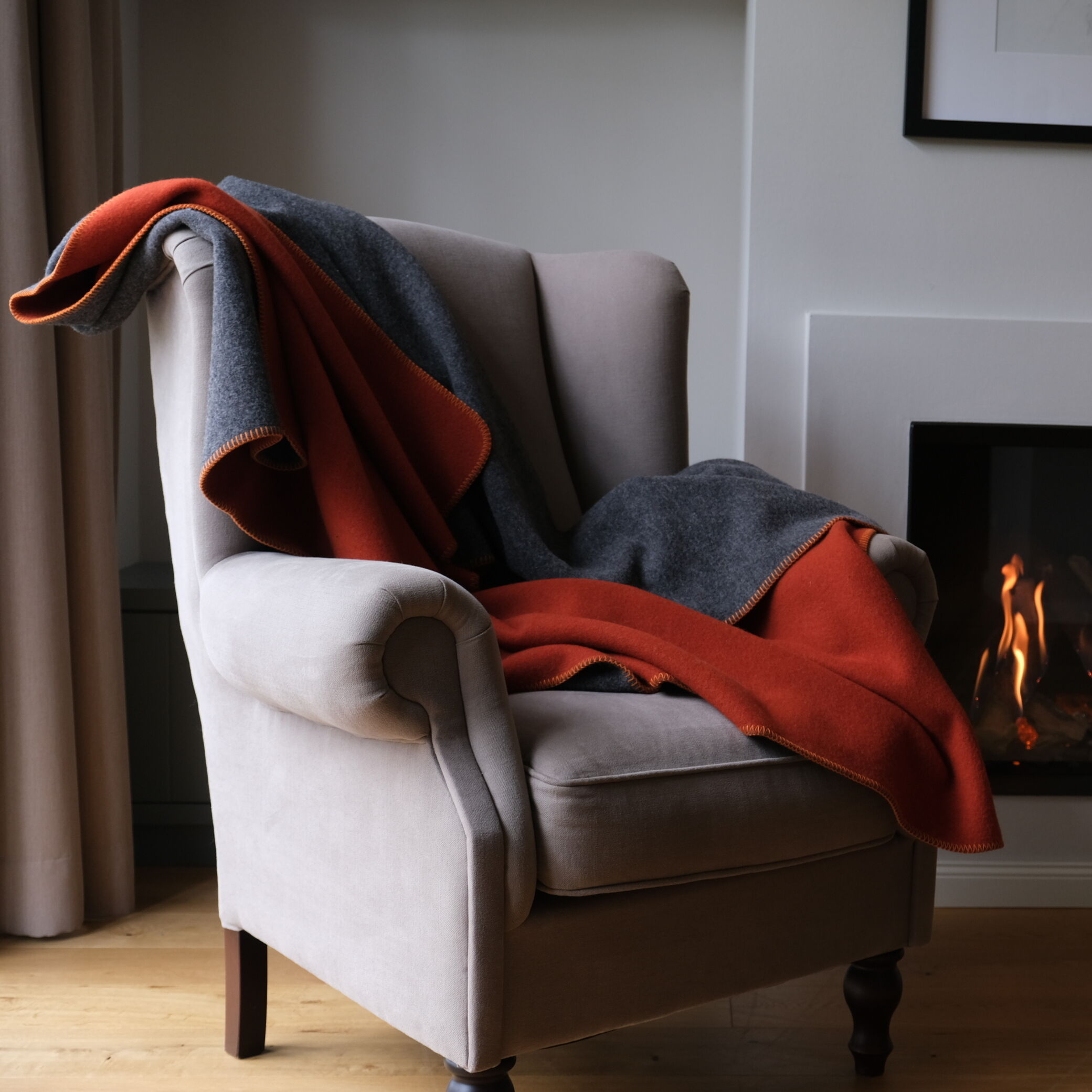 Eine Merinodecke in Orange und Grau liegt dekorativ auf einem beigefarbenen Sessel in einem stilvollen Wohnzimmer, neben einem brennenden Kamin. Die Szene vermittelt Gemütlichkeit und Wärme.